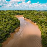 Vista aérea de uma embarcação navegando por um rio de águas barrentas em meio à floresta amazônica.