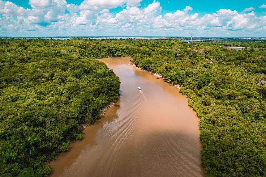 Vista aérea de uma embarcação navegando por um rio de águas barrentas em meio à floresta amazônica.