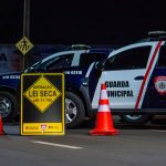 Agentes da Guarda Municipal realizam bloqueio policial noturno com viaturas e cones em uma via.