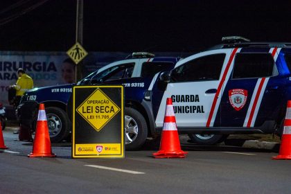 Agentes da Guarda Municipal realizam bloqueio policial noturno com viaturas e cones em uma via.