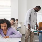 Grupo diverso de estudantes realiza uma prova em uma sala de aula moderna, sob supervisão de um professor.