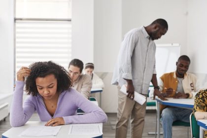 Grupo diverso de estudantes realiza uma prova em uma sala de aula moderna, sob supervisão de um professor.