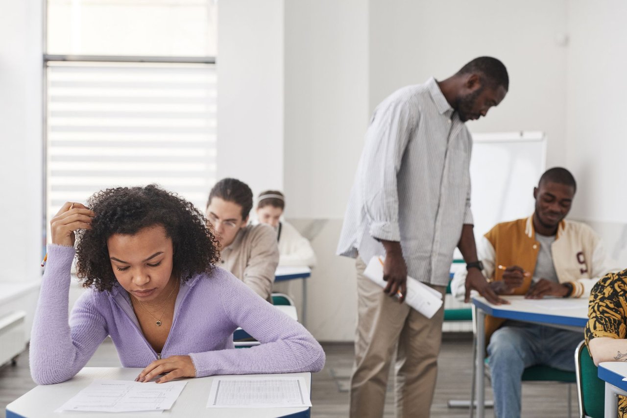 Grupo diverso de estudantes realiza uma prova em uma sala de aula moderna, sob supervisão de um professor.