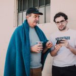 Voluntário ajuda um homem em situação de rua a usar o celular em um espaço ao ar livre.