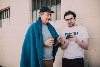 Voluntário ajuda um homem em situação de rua a usar o celular em um espaço ao ar livre.
