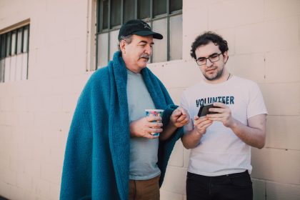 Voluntário ajuda um homem em situação de rua a usar o celular em um espaço ao ar livre.
