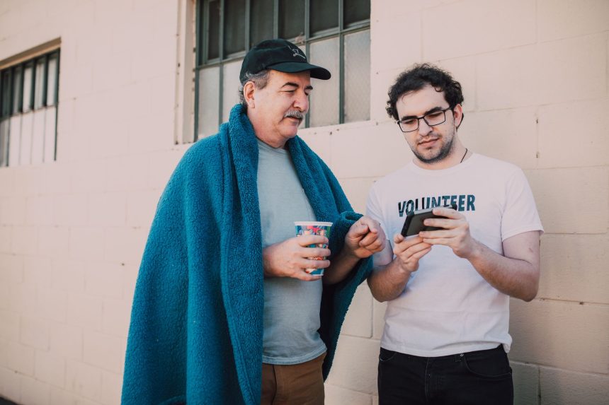 Voluntário ajuda um homem em situação de rua a usar o celular em um espaço ao ar livre.